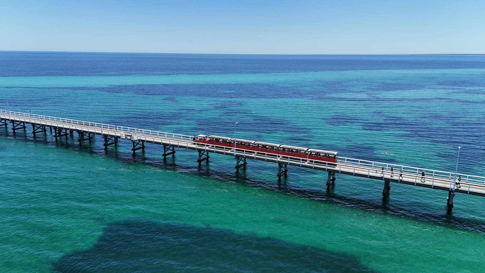 Aerial view of Busselton Jetty with a red train over vibrant turquoise waters in Western Australia.