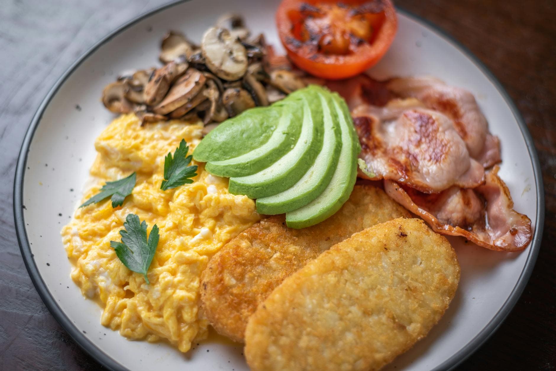 Scrambled eggs with avocado, bacon, mushrooms, and hash browns on a breakfast plate.