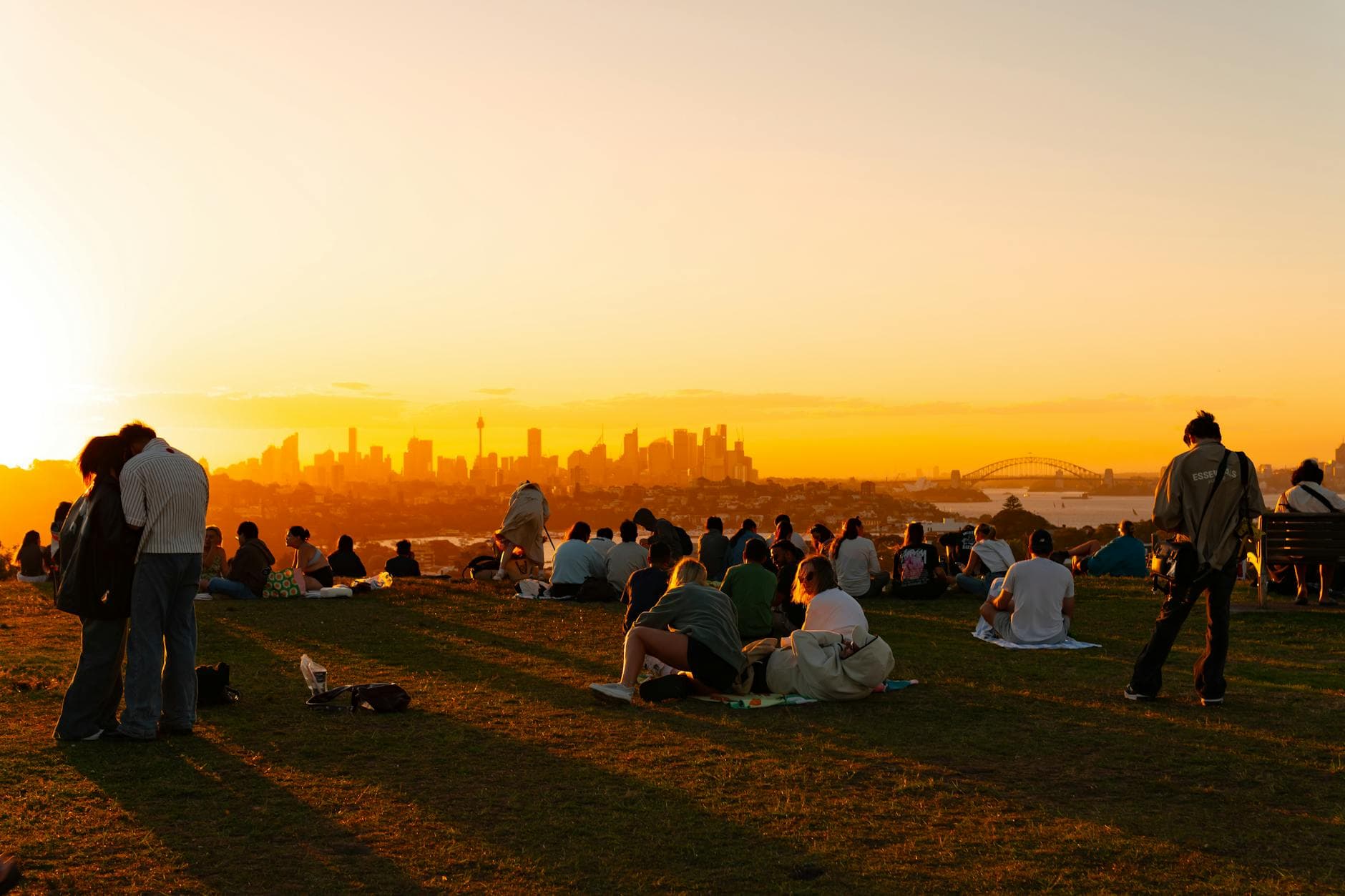 People enjoy a sunset view over Sydney skyline with the iconic Harbour Bridge visible.