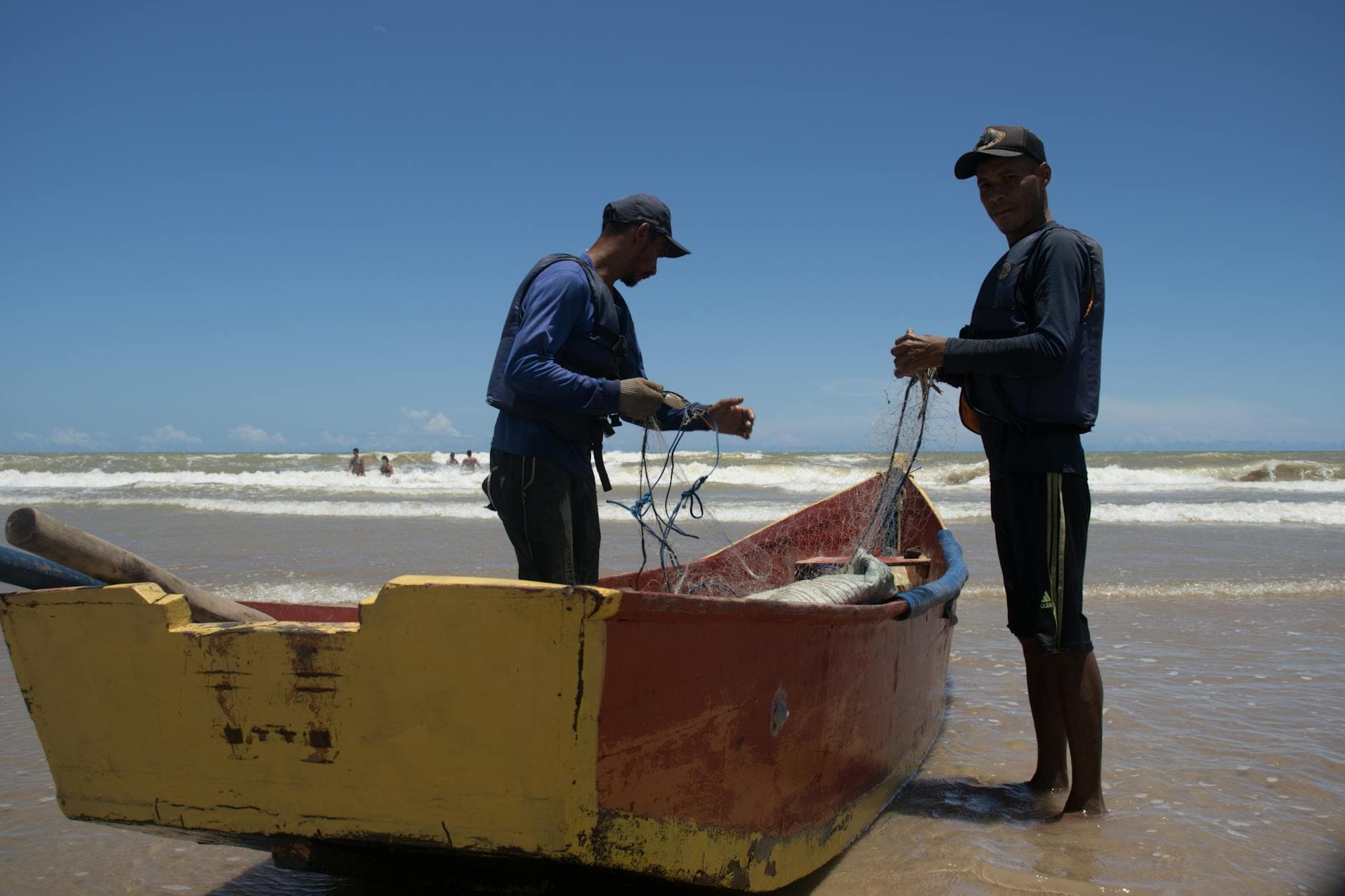 Two fishermen prepare their nets by a colorful boat on Itaúnas Beach, Brazil.