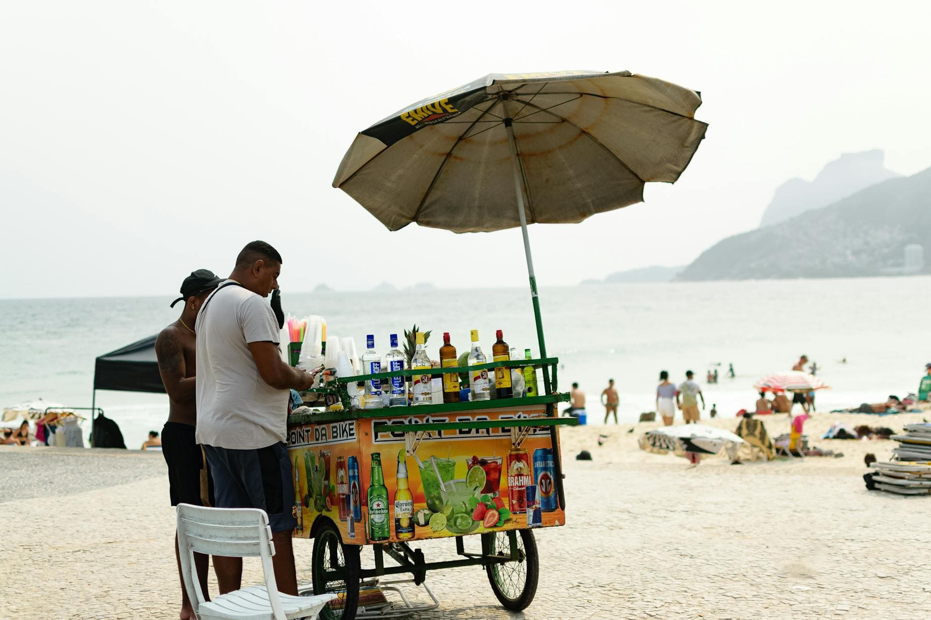Street vendor on Rio beach selling drinks under an umbrella with ocean backdrop.