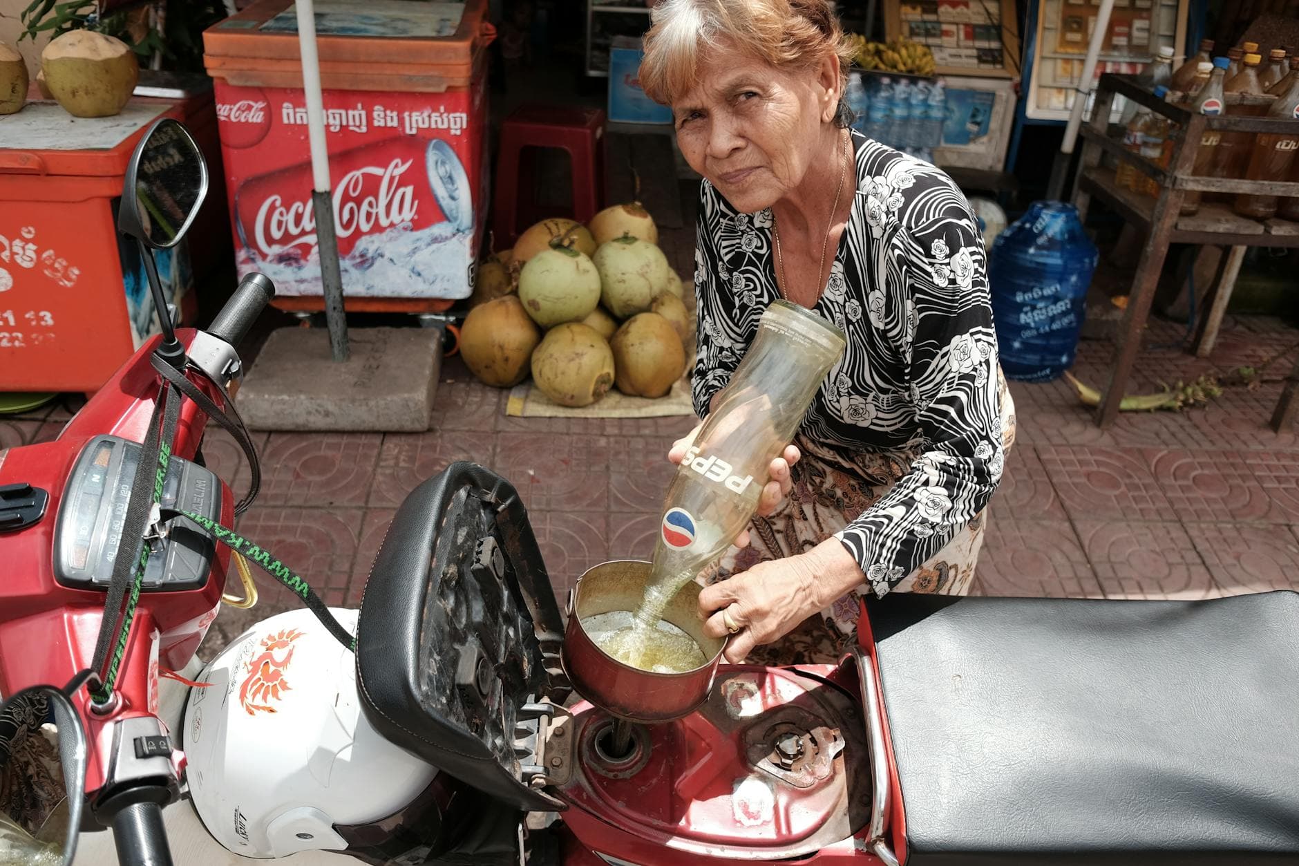 An elderly woman refills a glass bottle in a bustling Kampot street market.