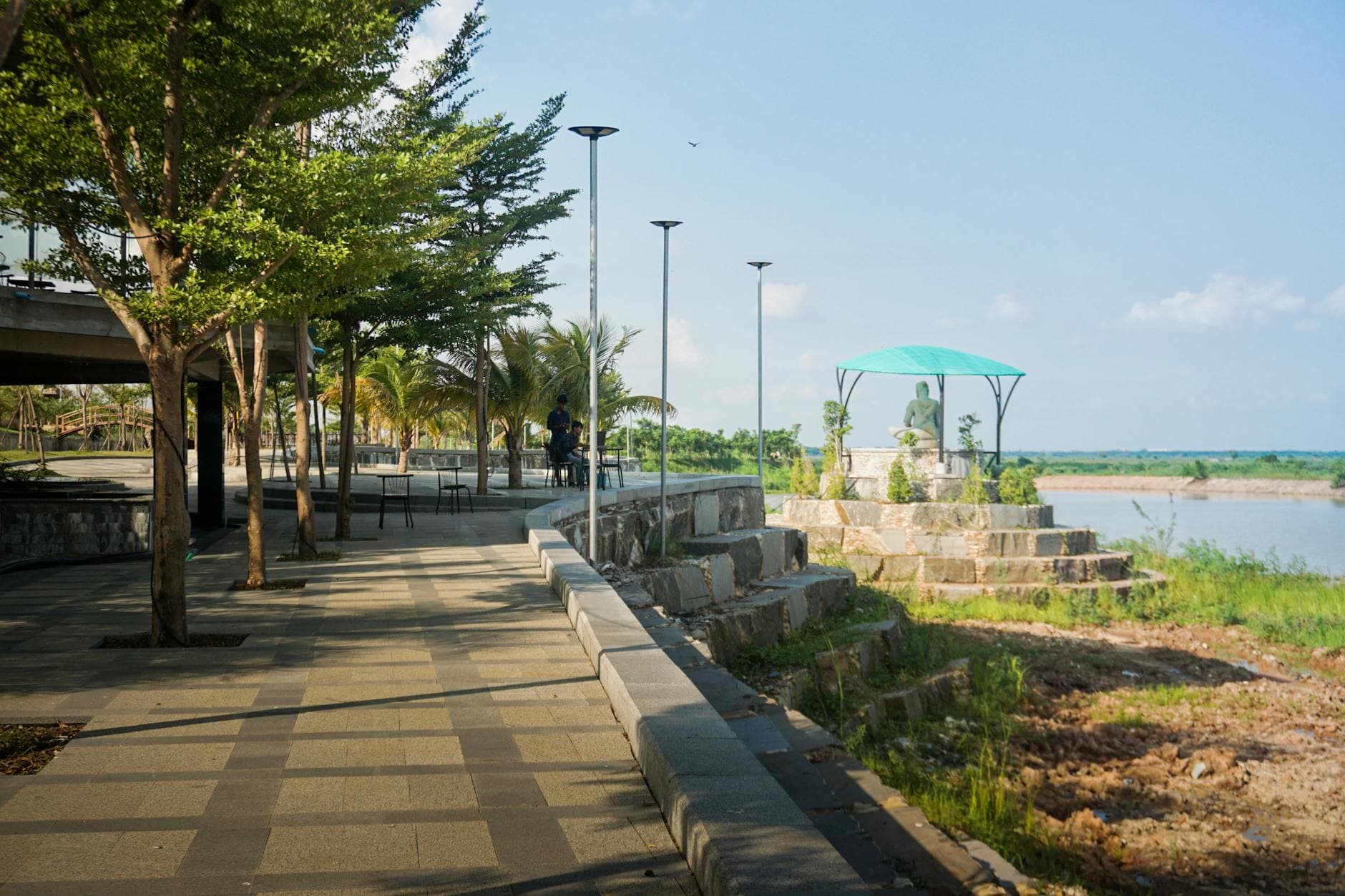Scenic riverside walkway with statue under canopy and lush trees in Cambodia.
