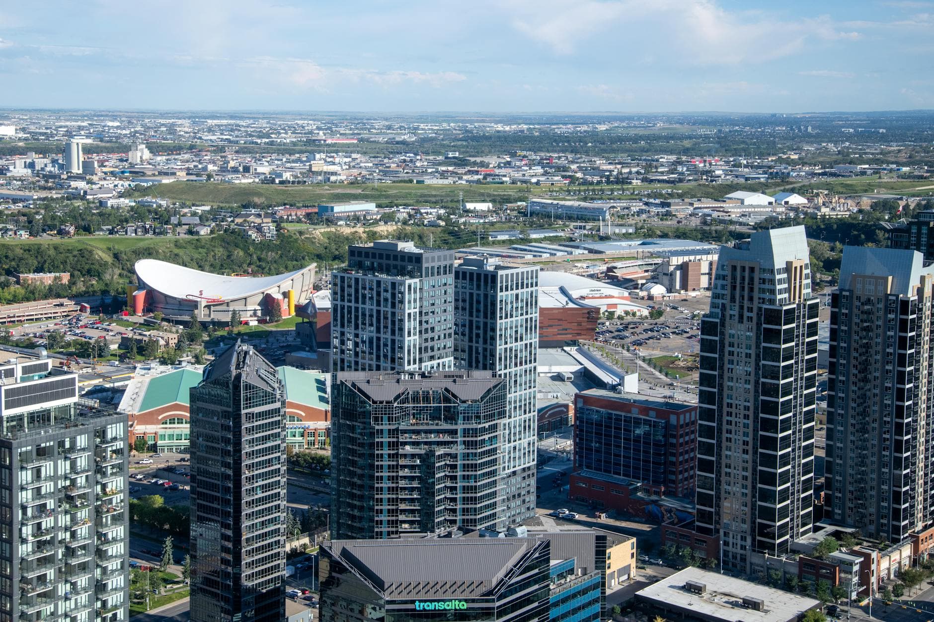 Aerial view of Calgary cityscape featuring modern skyscrapers and the iconic Saddledome under a clear blue sky.