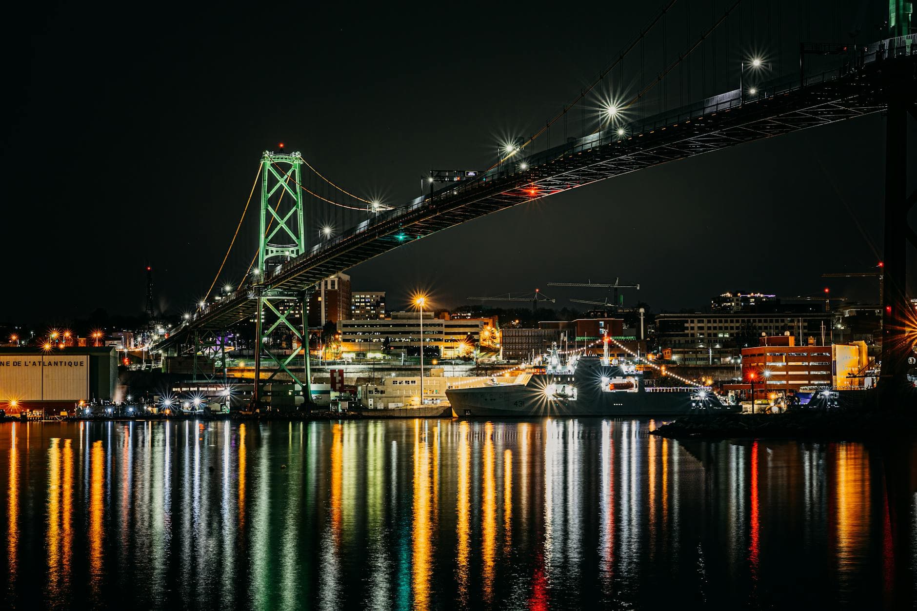 Night view of Halifax harbor featuring a bright suspension bridge with city lights reflecting on water.