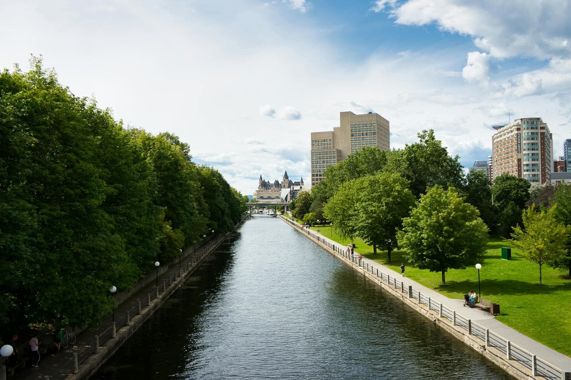 Tranquil urban scene featuring a canal, lush park, and city skyline under a blue sky.