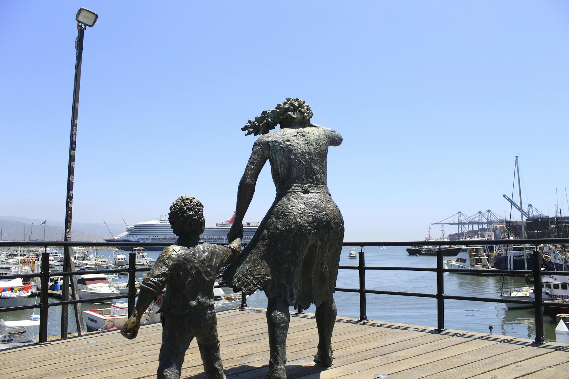 bronze statues of a woman and child overlooking a vibrant harbor with boats and a cruise ship.
