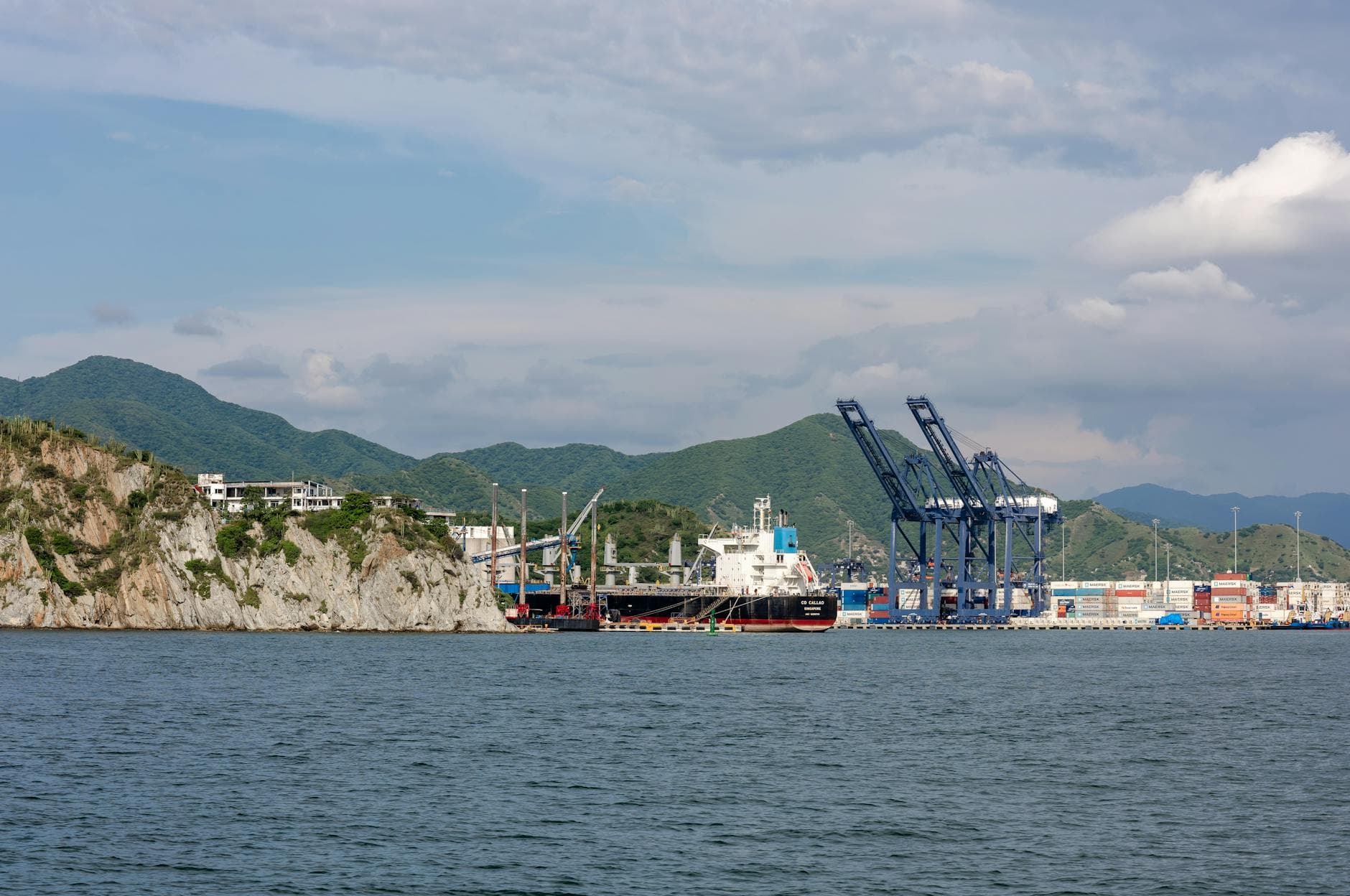 A vibrant coastal trading port with cranes, surrounded by sea cliffs and mountains under a bright sky.
