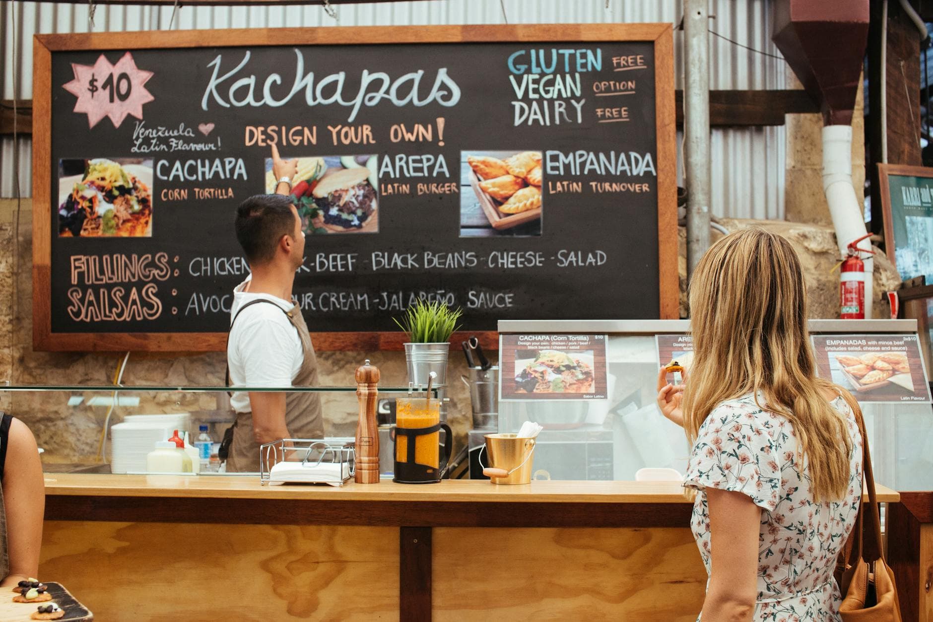 Customers at a Venezuelan snack bar choosing customizable food options from a colorful menu board.