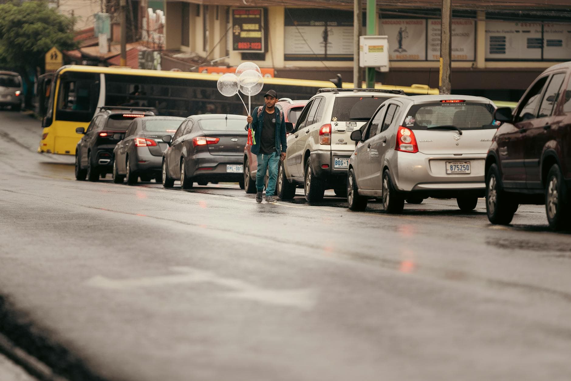 A street vendor in Alajuela, Costa Rica, selling balloons amidst traffic. Captured on a rainy day.