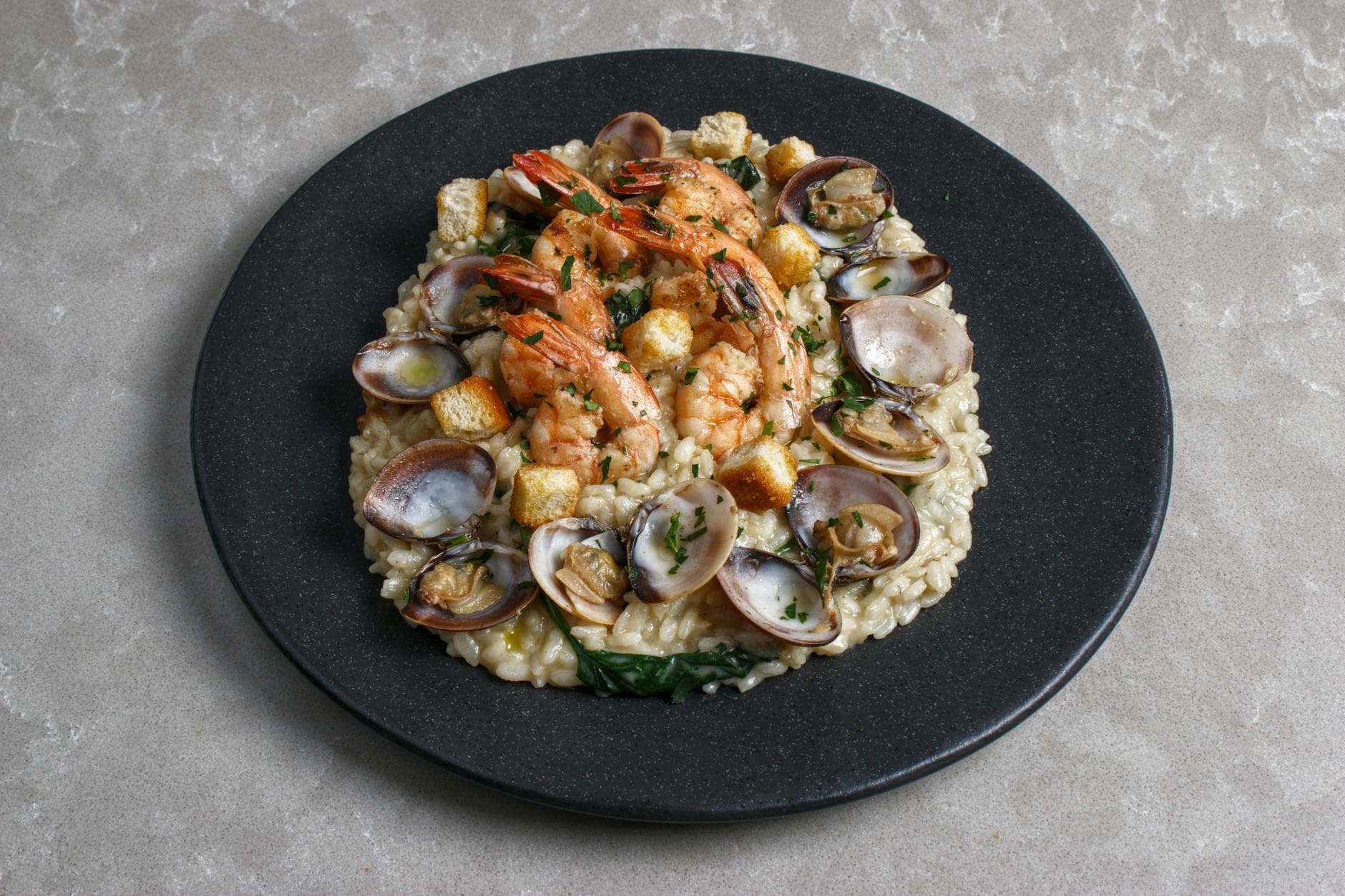 A gourmet seafood risotto topped with succulent shrimp and clams served on a black plate.