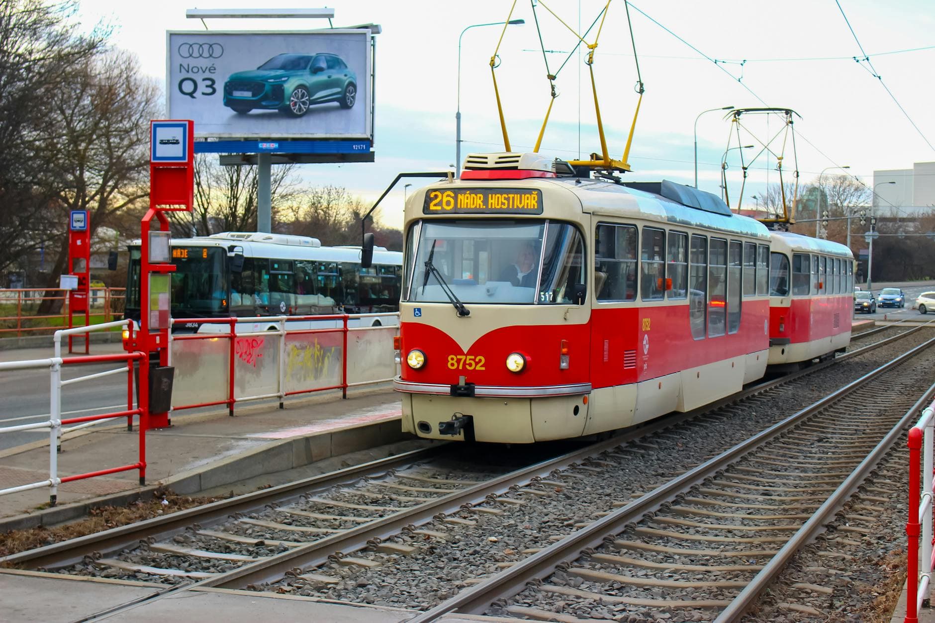 A red tram captured in motion on the streets of Prague, Czechia. Billboard and trees in the background.