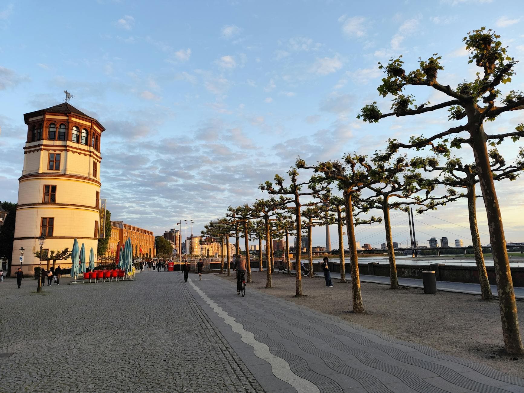 Scenic view of Düsseldorf's riverside promenade with tower and trees at sunset.