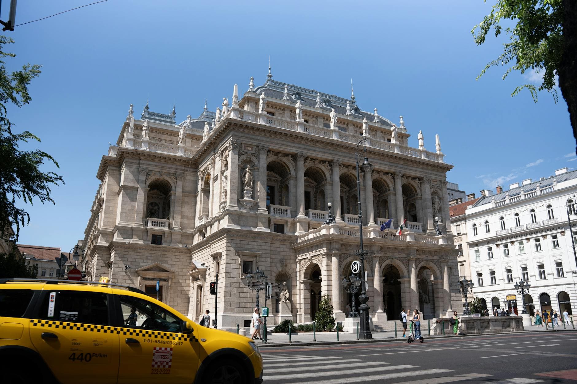 Street view of Budapest's Opera House with a yellow taxi passing by, iconic architecture.