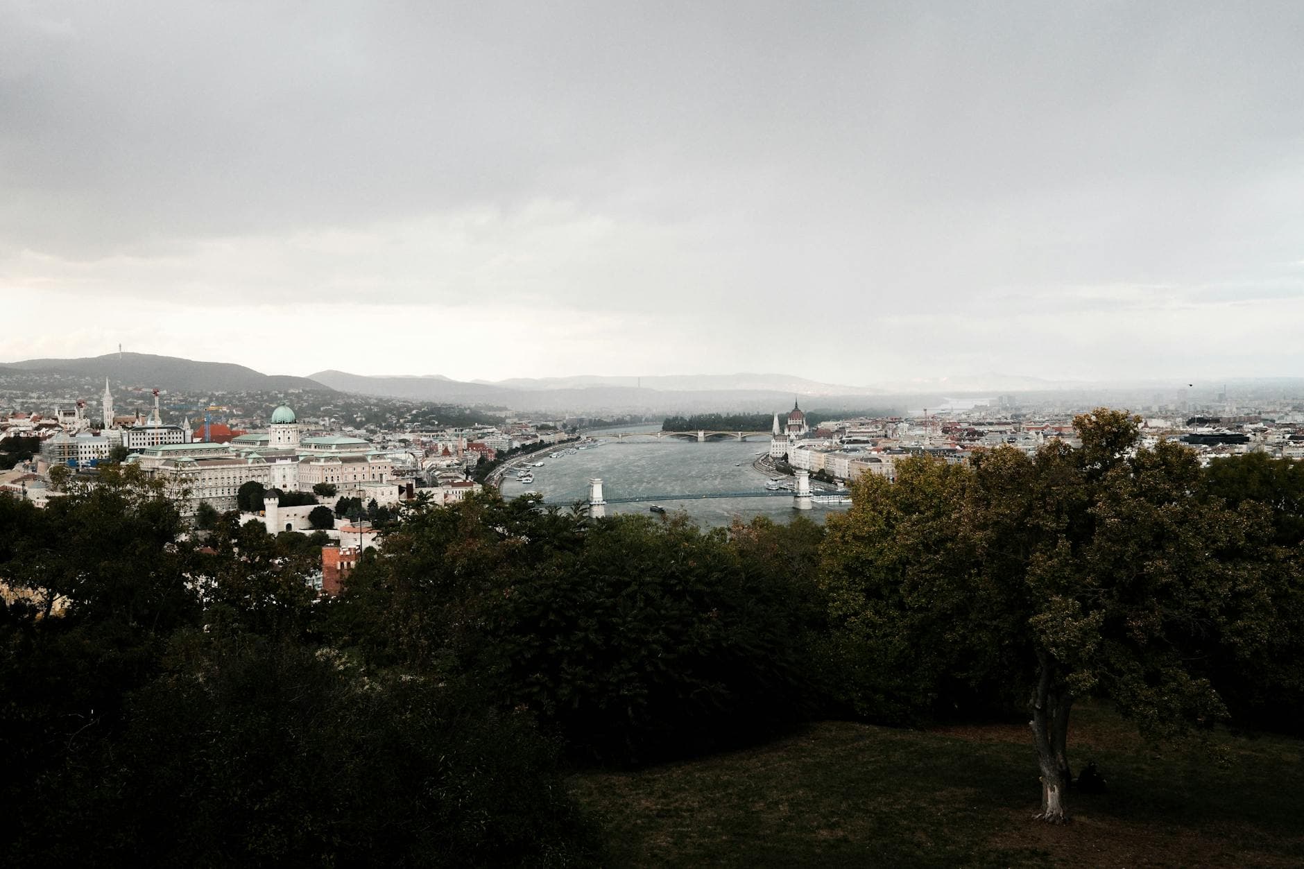 Scenic view of Budapest's cityscape with the Danube River and iconic landmarks under a cloudy sky.