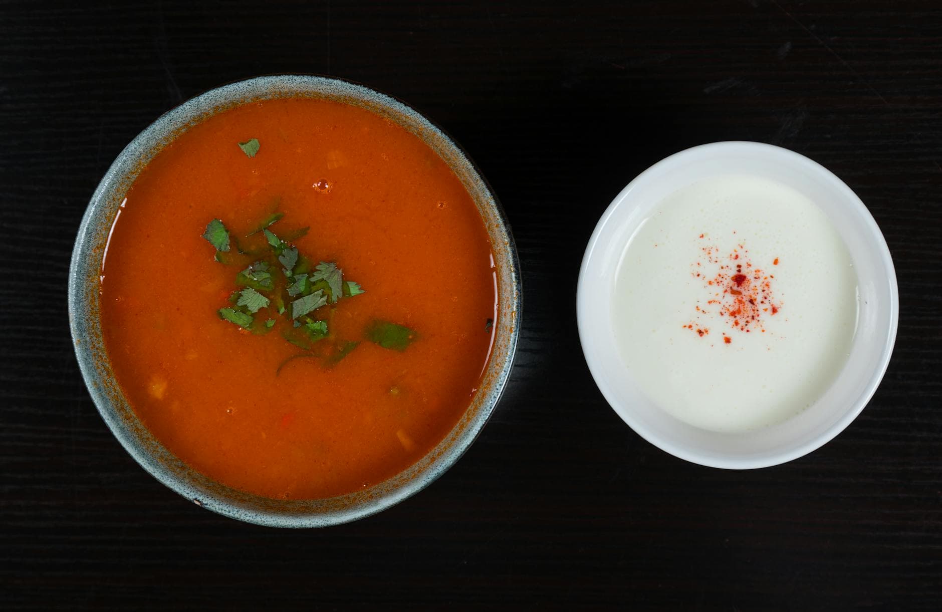 A vibrant tomato soup garnished with cilantro beside a bowl of creamy yogurt with paprika.