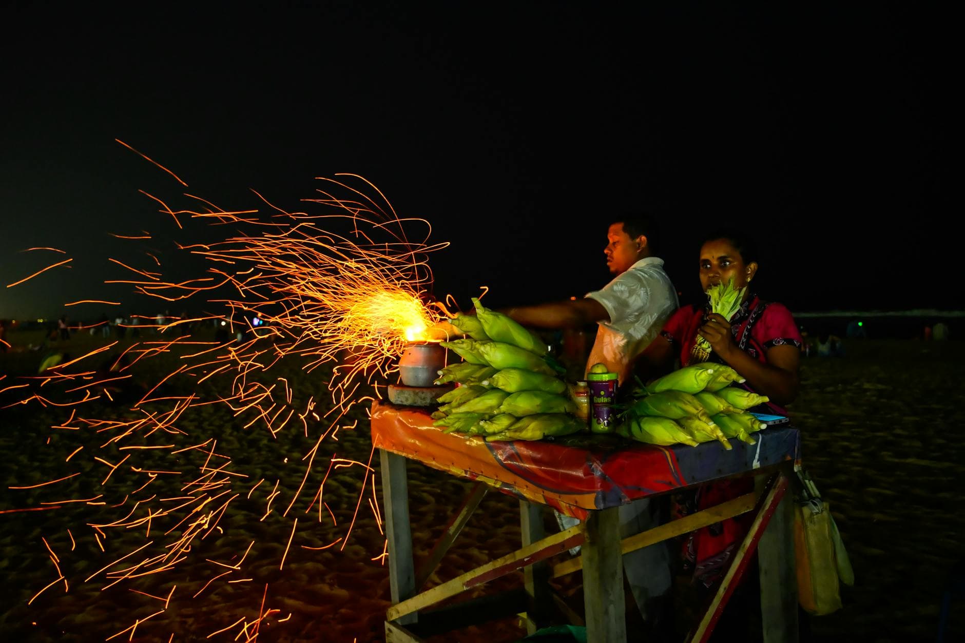 Dynamic nighttime scene of a street vendor grilling corn on Chennai beach, sparks flying.
