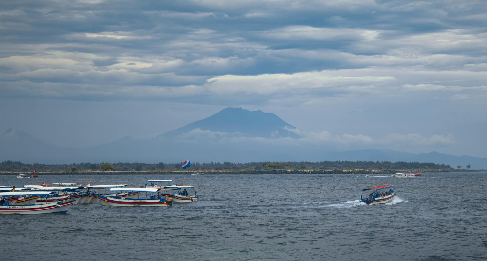 Colorful boats on a calm sea with a majestic mountain and cloudy sky in the background, creating a serene landscape.
