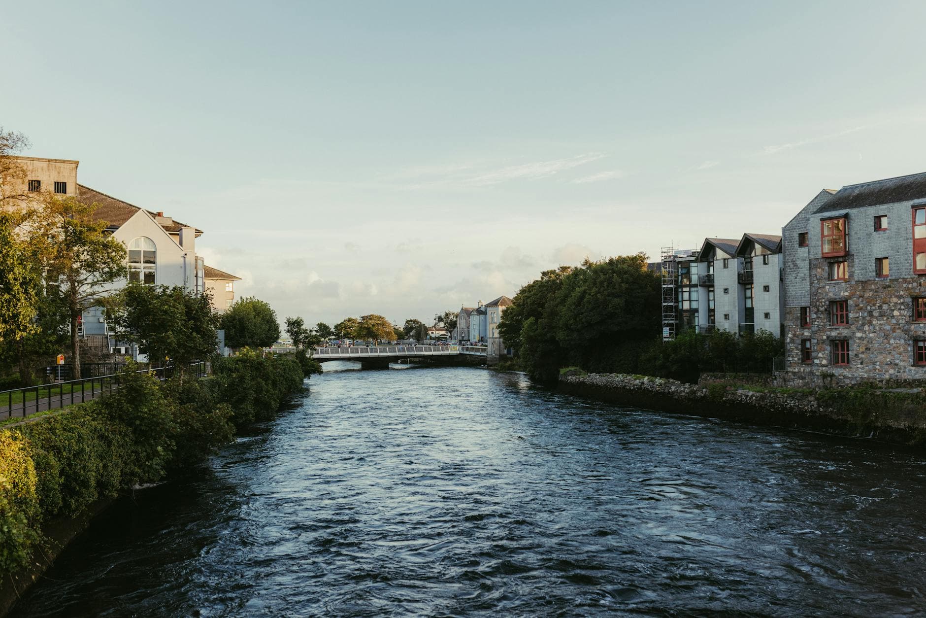 Tranquil river scene in Galway with historic stone buildings and lush greenery at sunset.