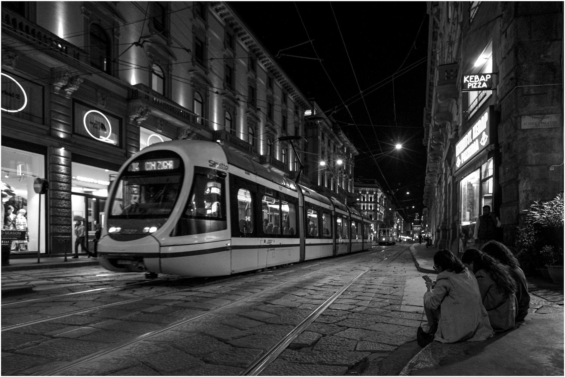 Black and white photo capturing a nighttime tram in urban Milano with people sitting nearby.
