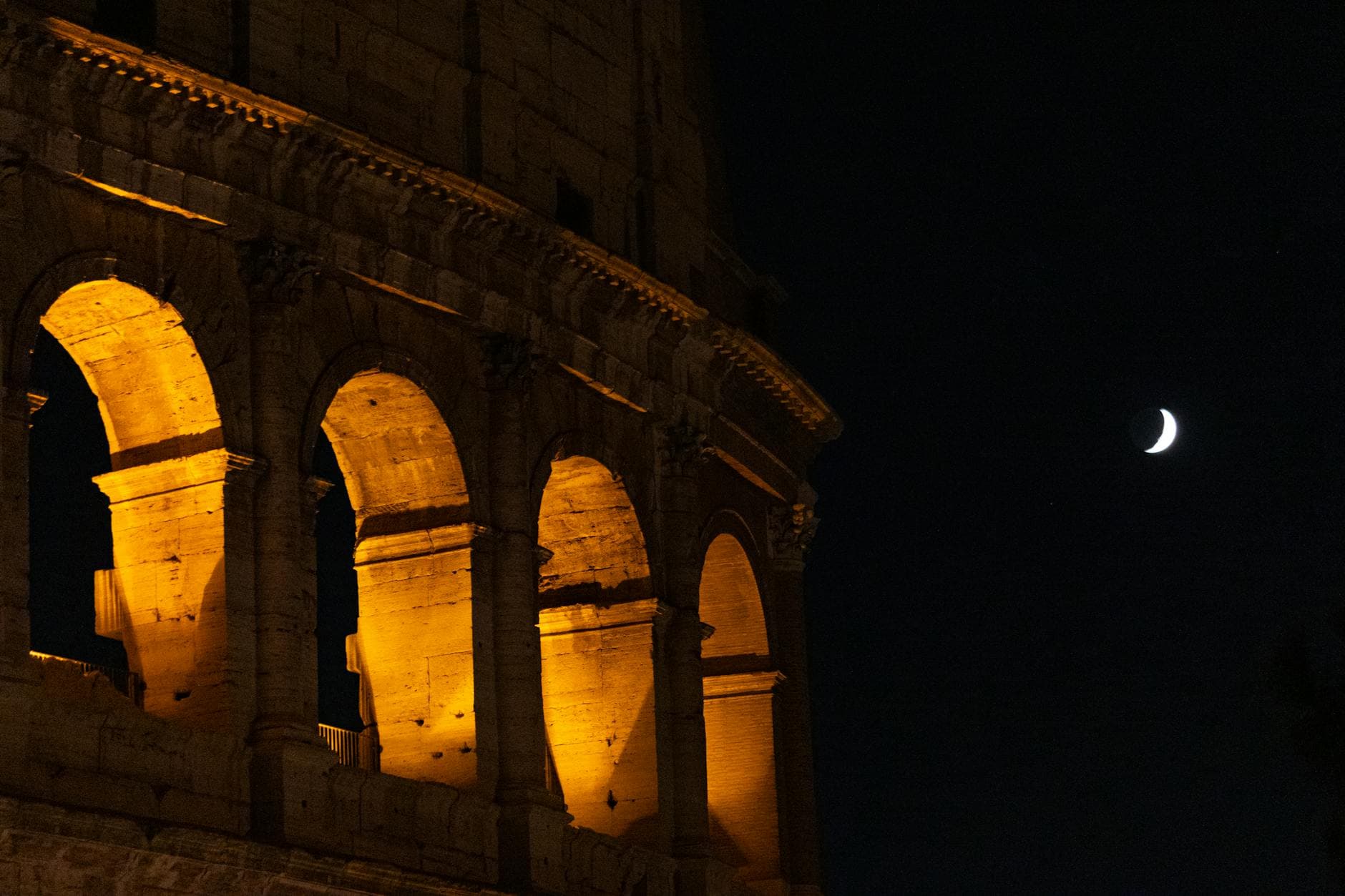 Beautiful nighttime view of the Colosseum in Rome with a crescent moon in the sky.
