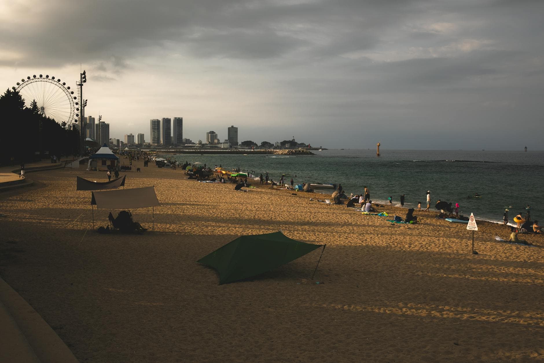 A tranquil beach scene shows a ferris wheel and city skyline under a cloudy dusk sky.