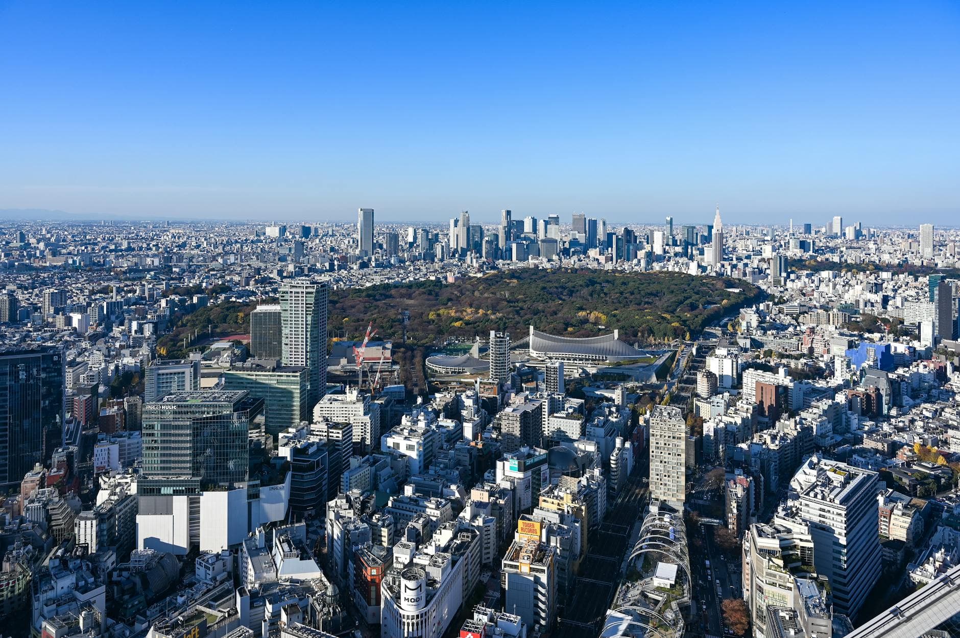 Aerial view of Tokyo cityscape featuring Shinjuku Park surrounded by skyscrapers under a clear blue sky.