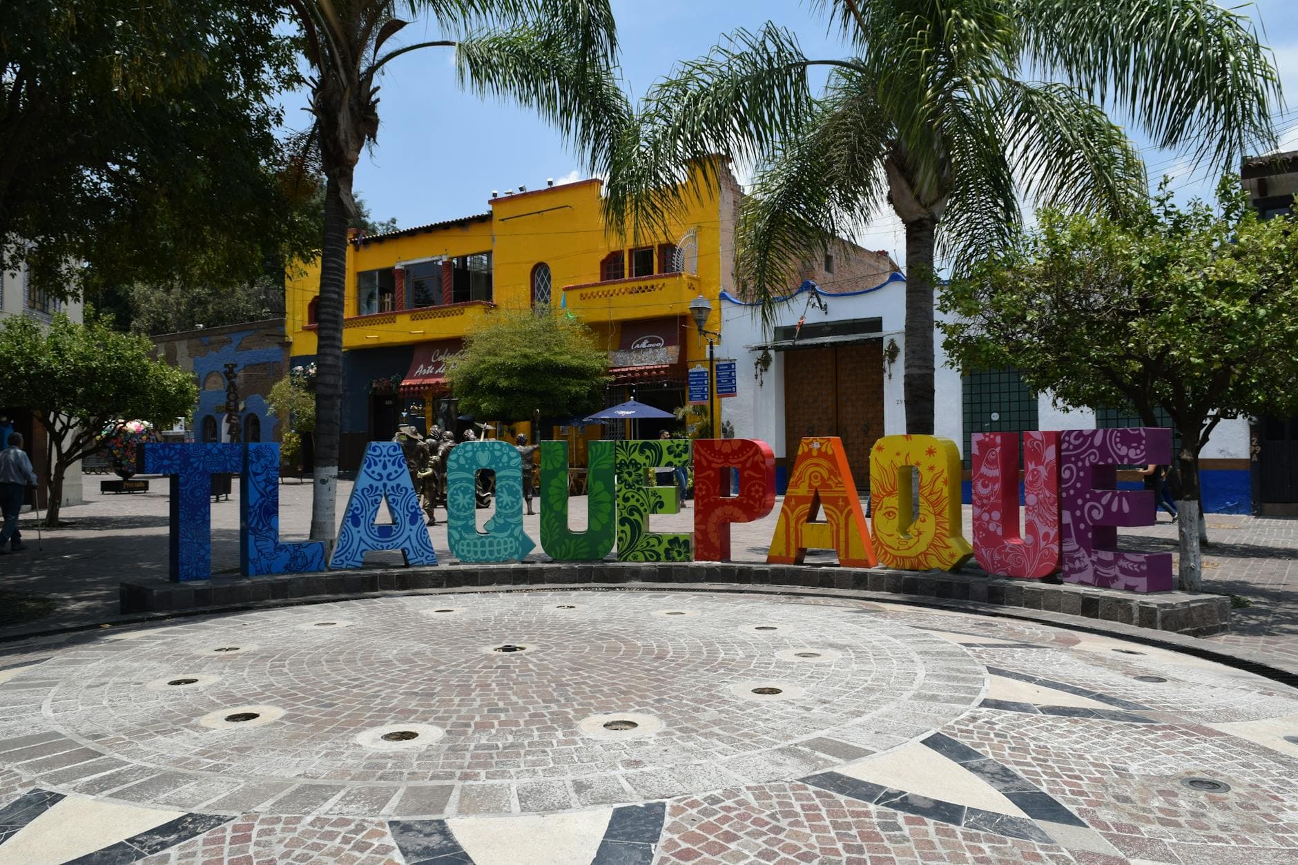 Vibrant Tlaquepaque sign in a lively Mexican plaza with palm trees and buildings.