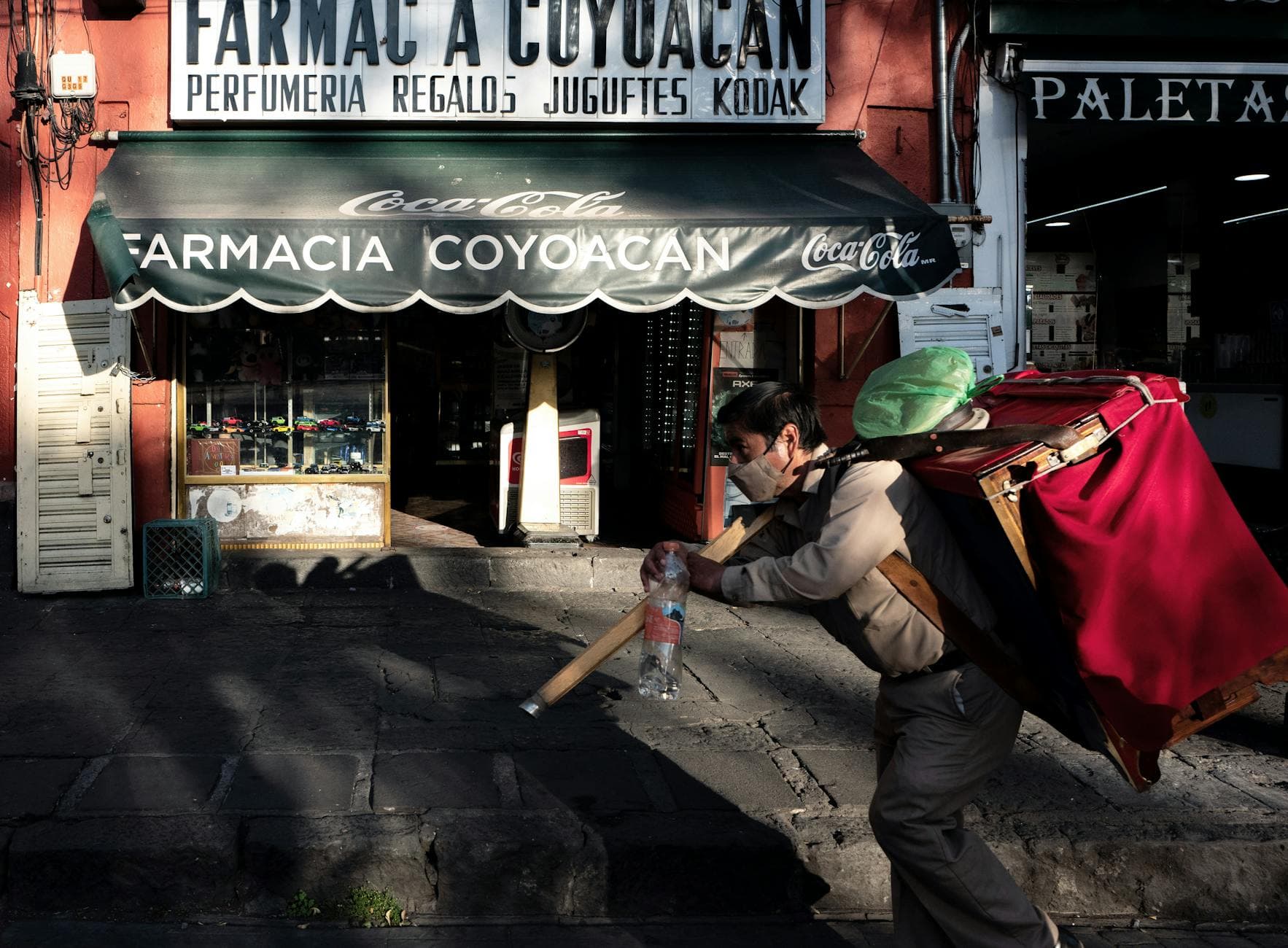 Man carrying goods past a pharmacy in Coyoacán, Mexico City, capturing vibrant urban life.