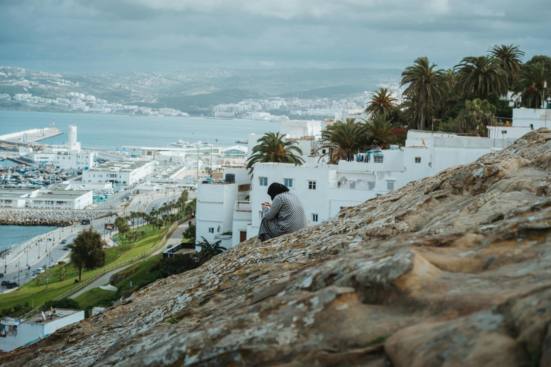 Woman sitting on hill overlooking Tangier port, capturing urban landscape.