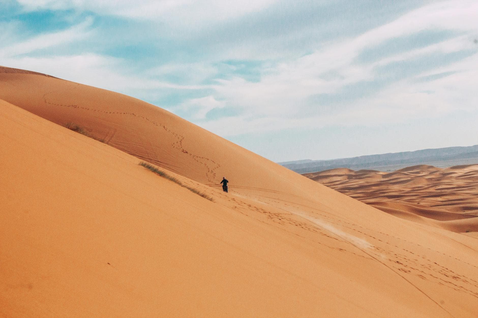 A lone traveler walks across the vast sand dunes of Merzouga, Morocco, under a clear sky.