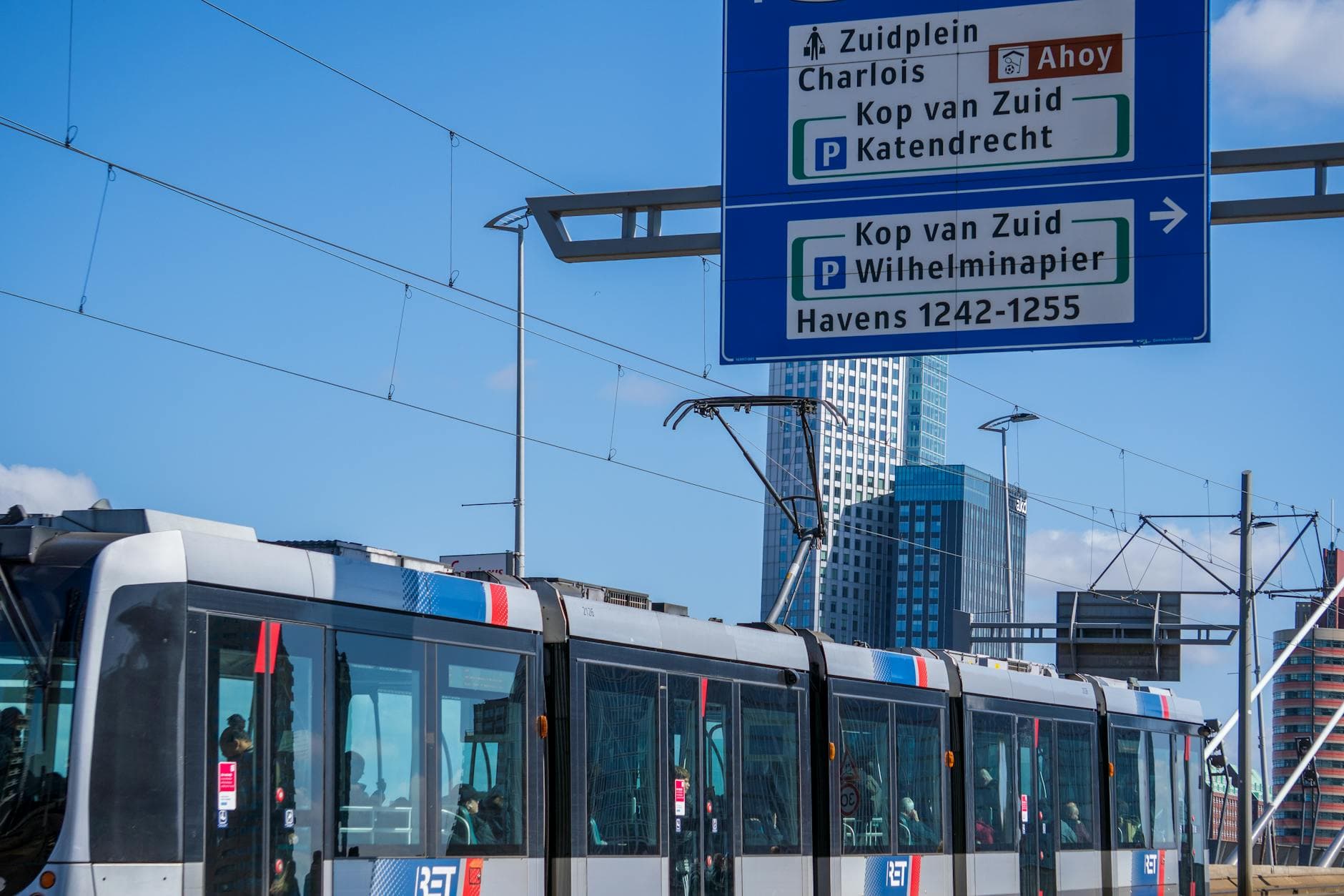 A modern tram passing by high-rise buildings in Rotterdam under a clear blue sky.