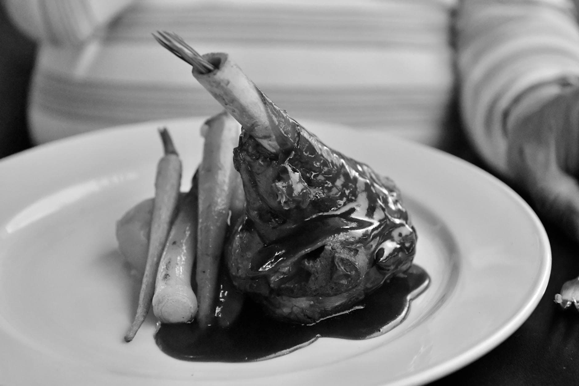 Black and white photo of lamb shank with roasted carrots and potatoes in sauce.