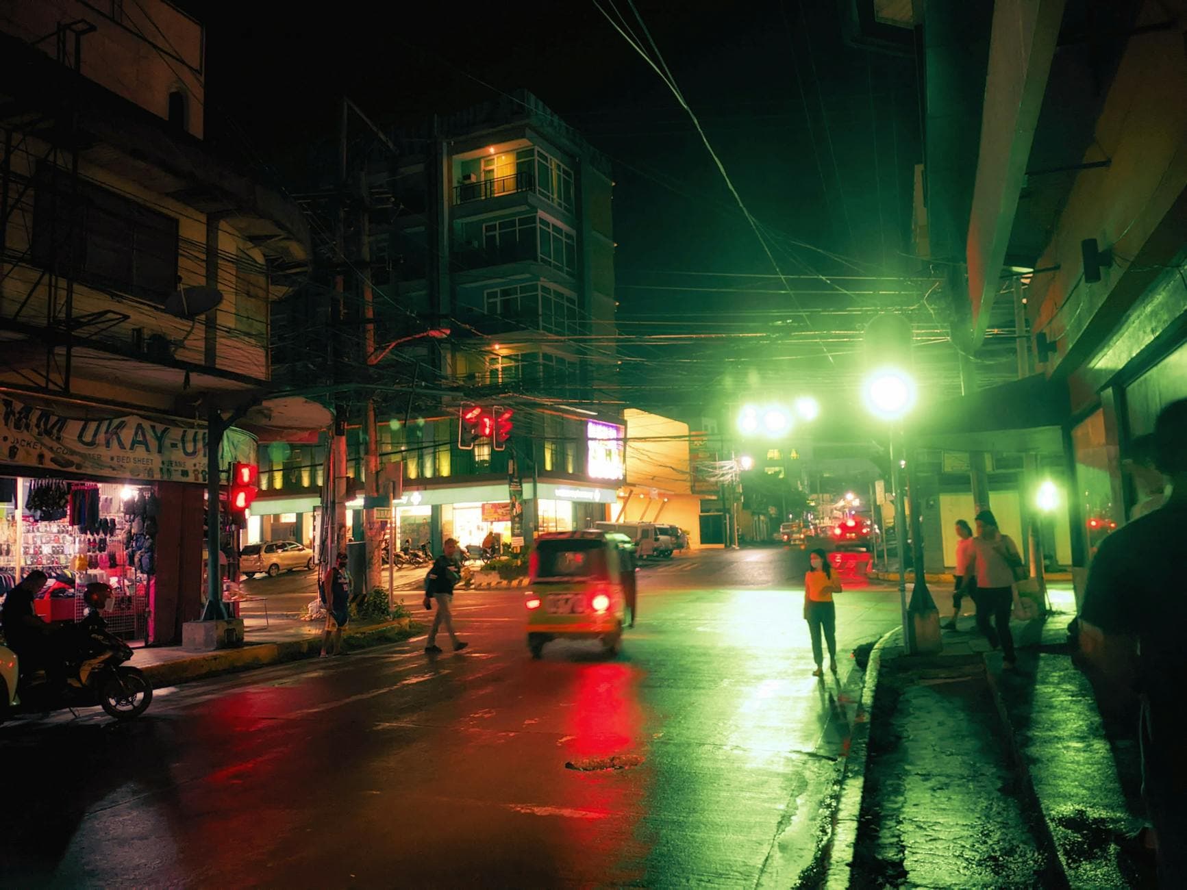 Colorful city nightlife on a bustling street corner with people and vehicles under green lights.