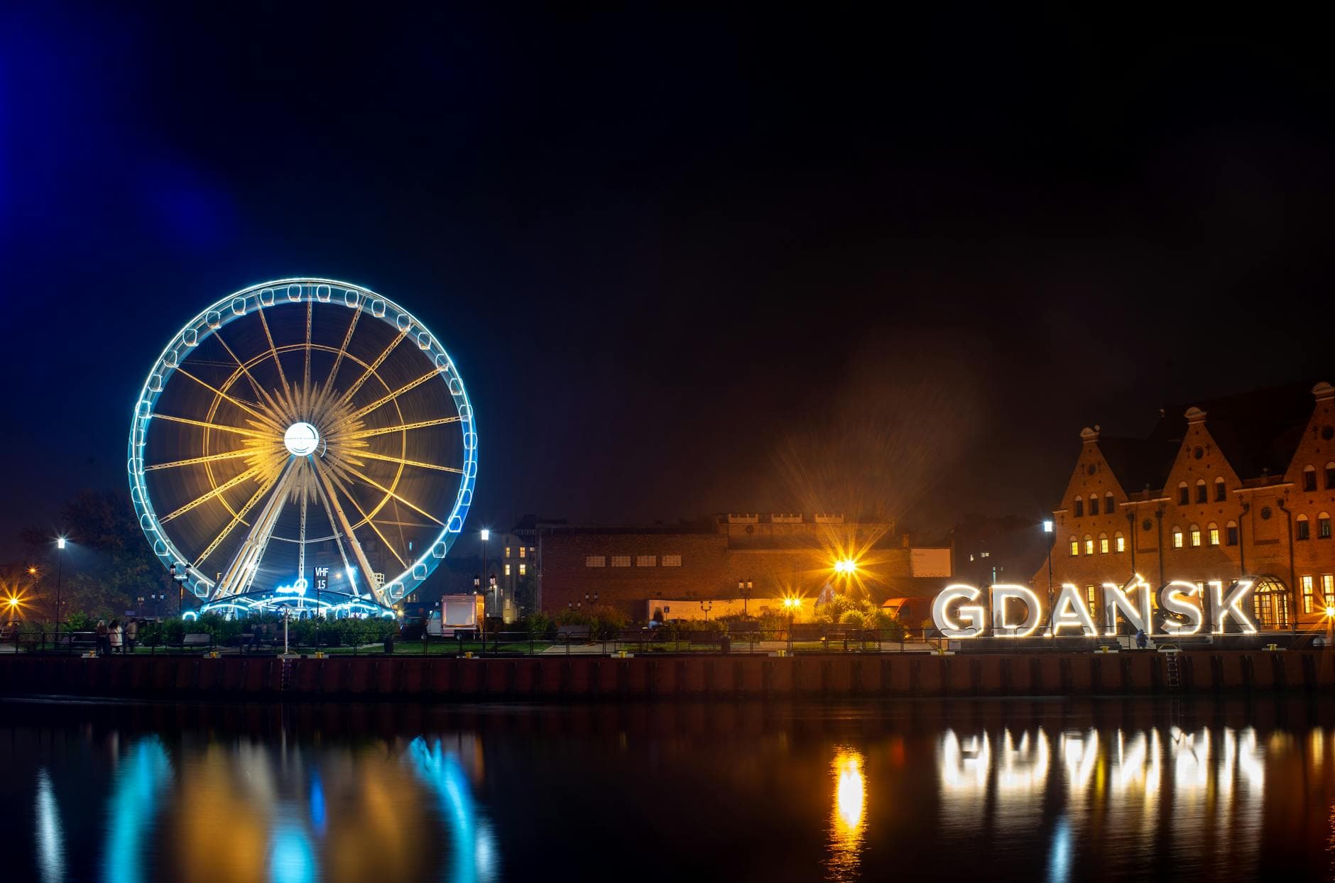 Night view of the brightly lit Ferris wheel and waterfront in Gdansk, Poland, reflecting in the water.