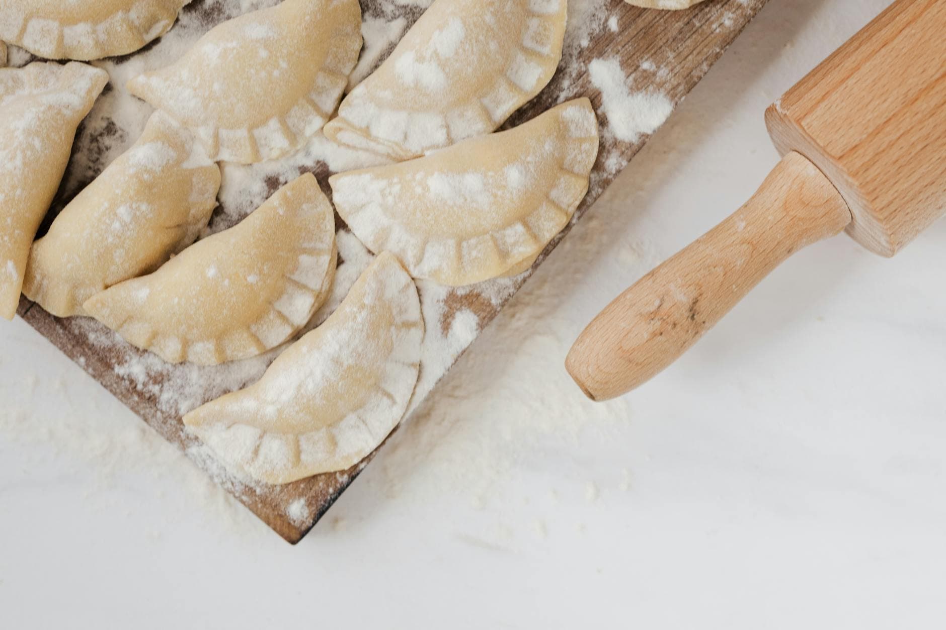 Close-up of uncooked pierogi on a floured wooden tray with a rolling pin, ready to cook.