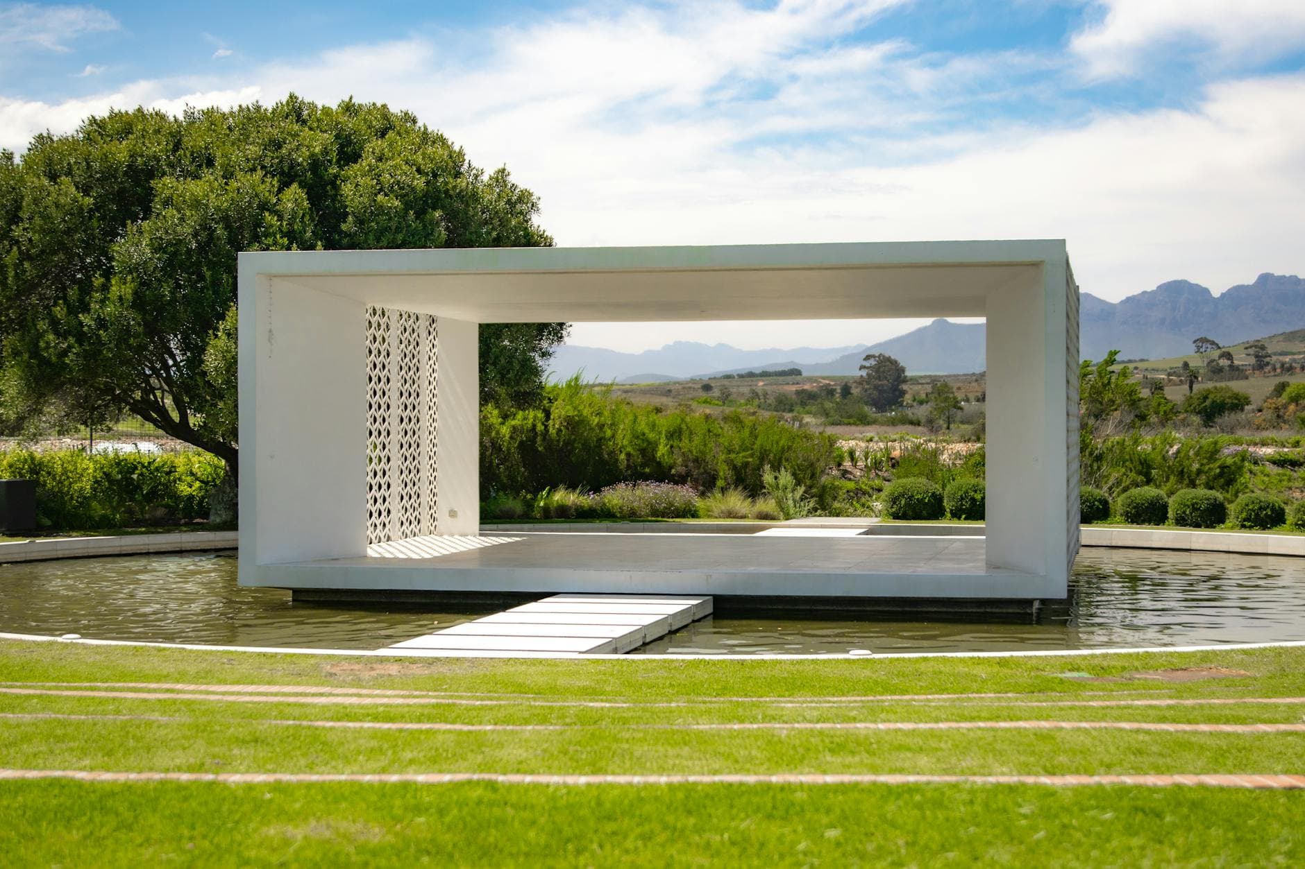 A contemporary structure by a pond in a lush vineyard setting with mountain views.