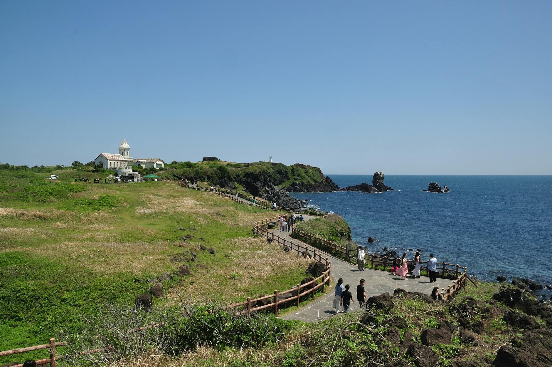 A picturesque view of Jeju Island's coastline with a walking path and a church in the background.