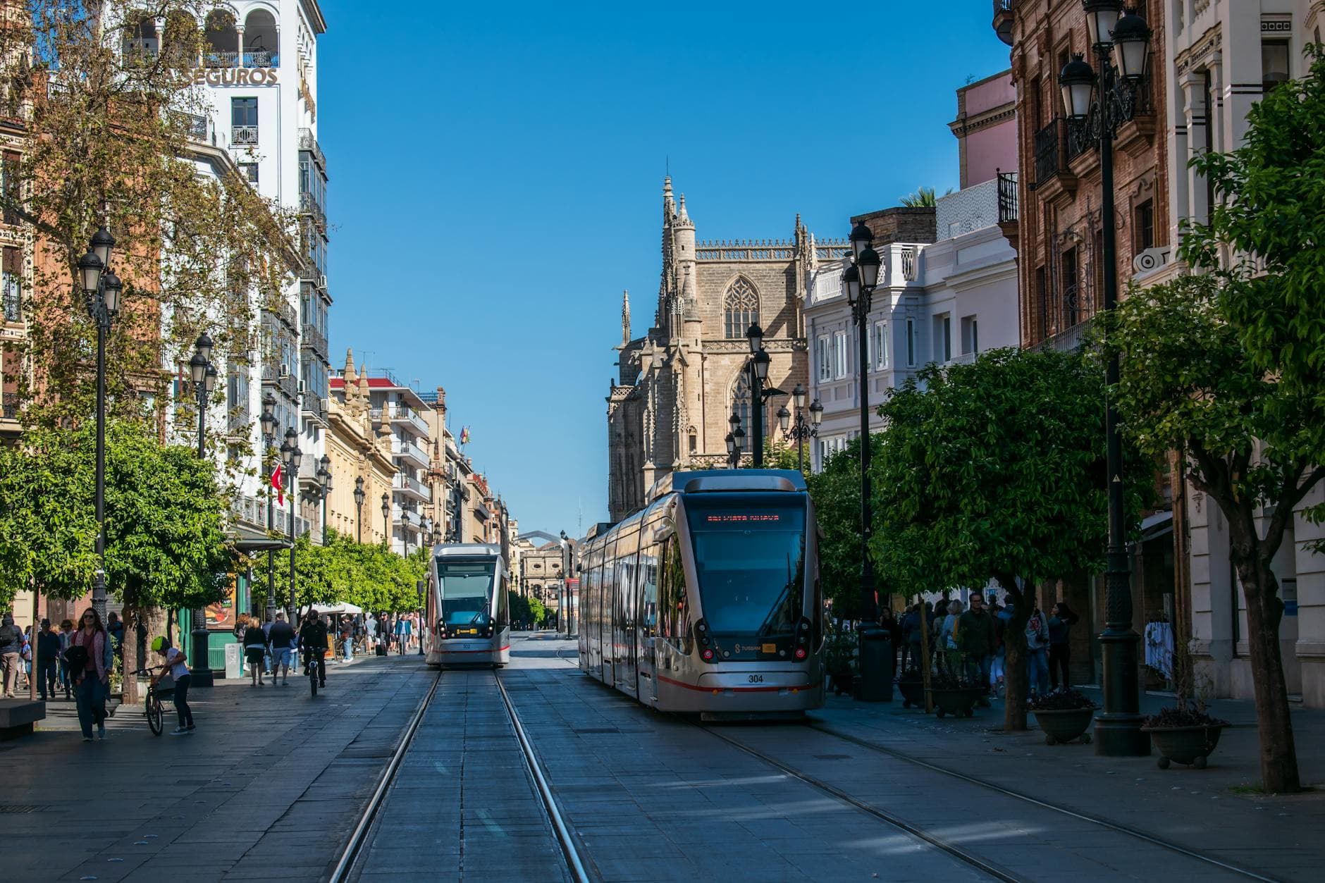 Trams traverse Avenida de la Constitución in Seville, showcasing urban architecture and street life.