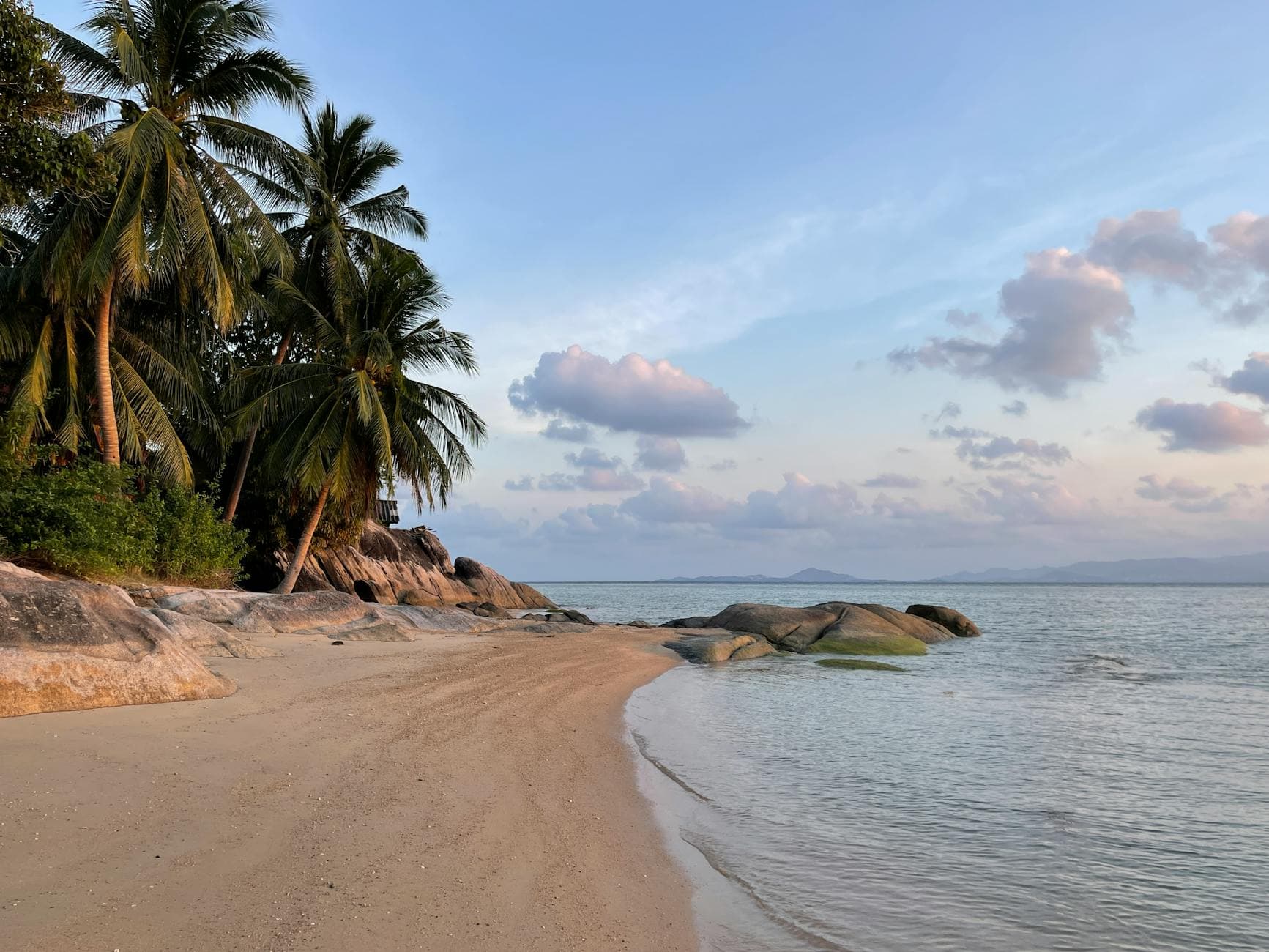 Peaceful tropical beach scene with palm trees, sand, and rocks under a blue sky.