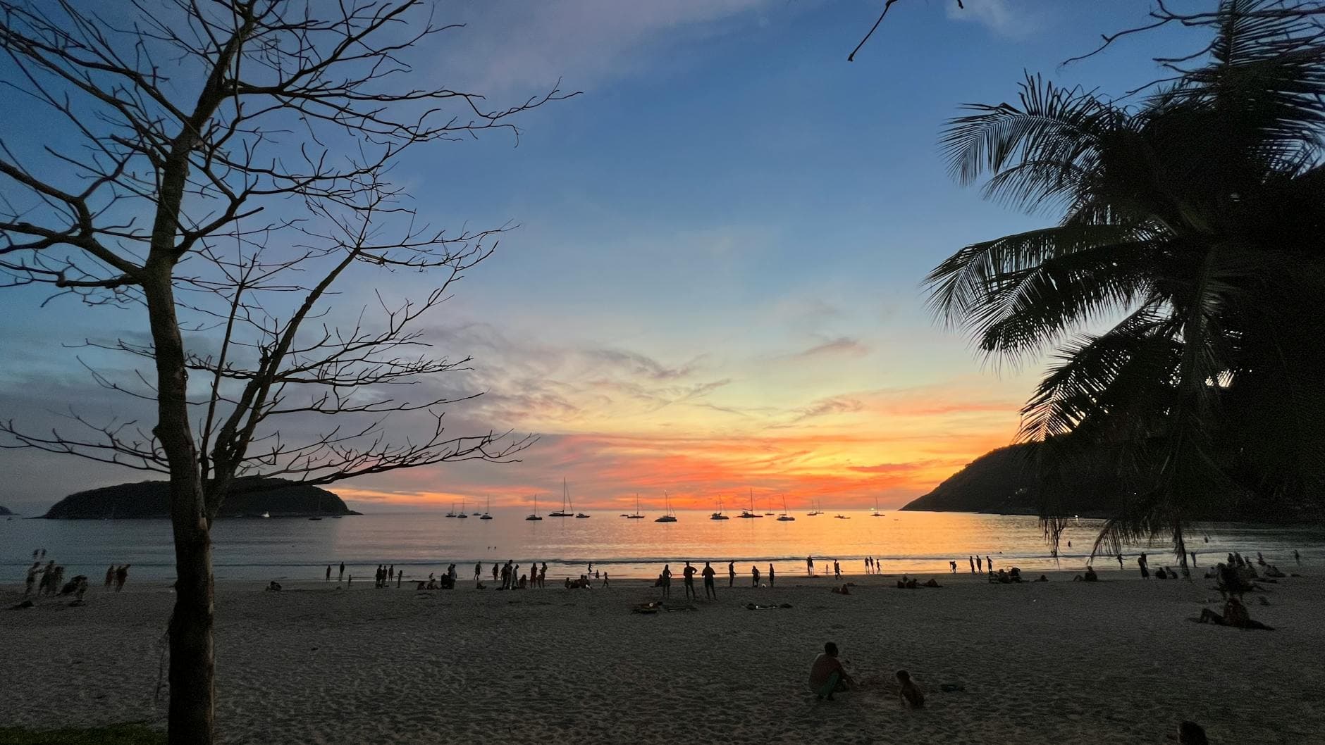 Beautiful sunset over a busy beach in Phuket, Thailand, with boats on the horizon and palm trees.