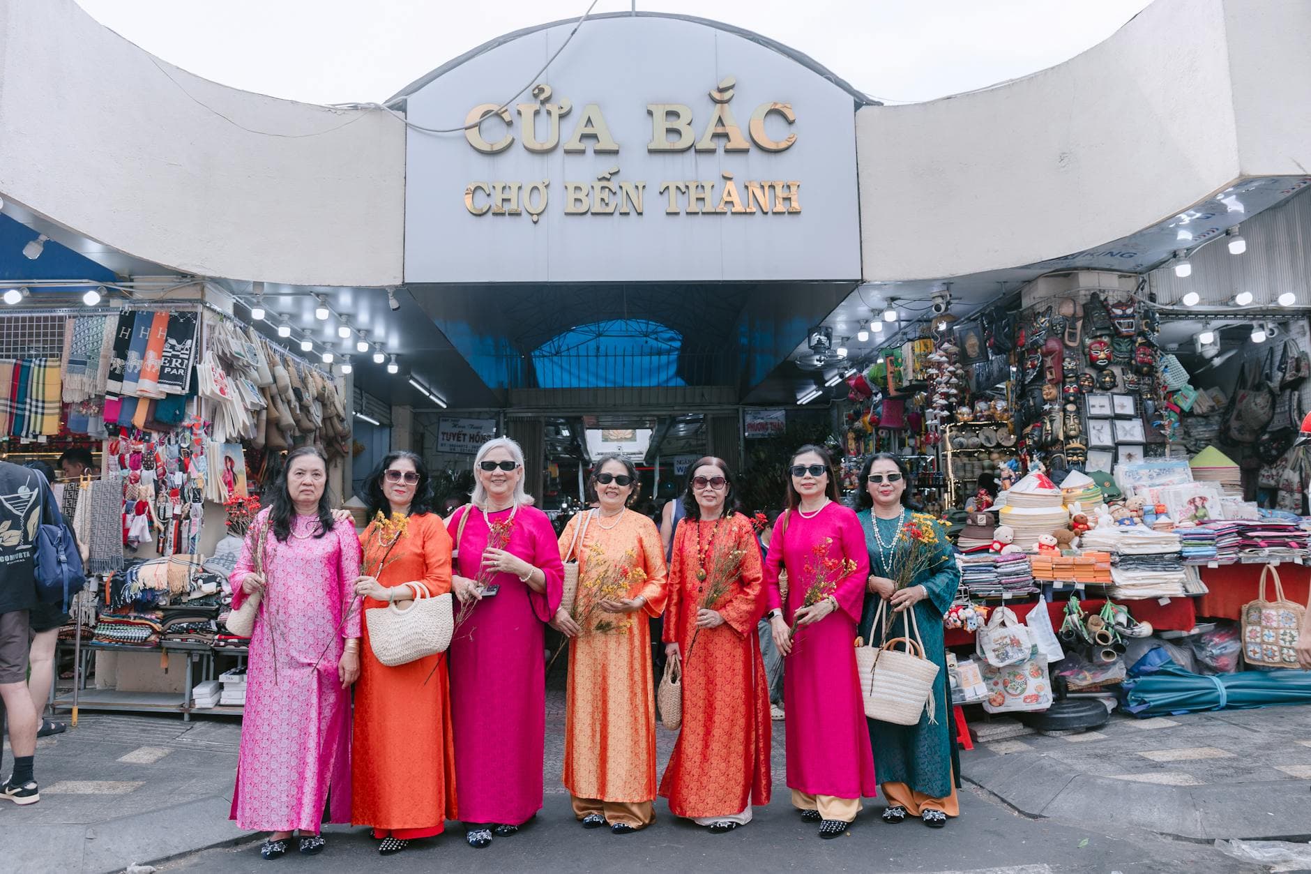 Group of women in vibrant traditional dresses posing at Ben Thanh Market entrance.